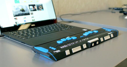 The image shows a refreshable Braille display connected to a laptop, allowing visually impaired users to navigate digital content via tactile Braille cells and buttons.