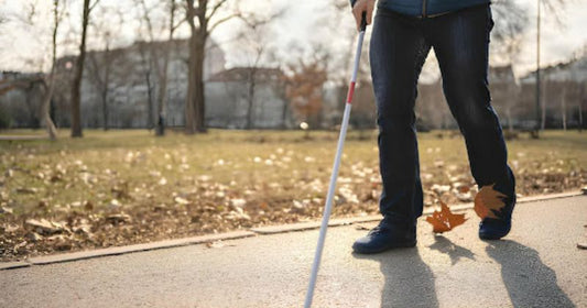 A visually impaired person walks along a park path using a white cane on a sunny autumn day.