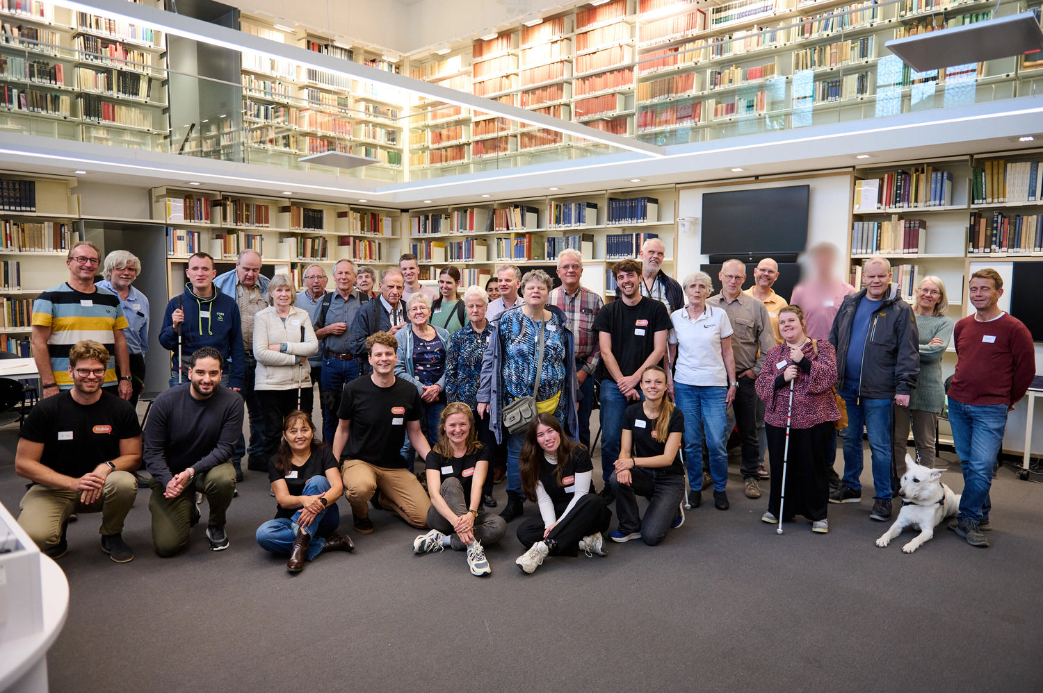 "A large group of blind and visually impaired people, along with organizers, pose together in a library space filled with bookshelves. Some participants hold white canes or are accompanied by guide dogs. The photo was taken during a Hable testing day. Everyone is smiling, creating a warm and welcoming atmosphere.