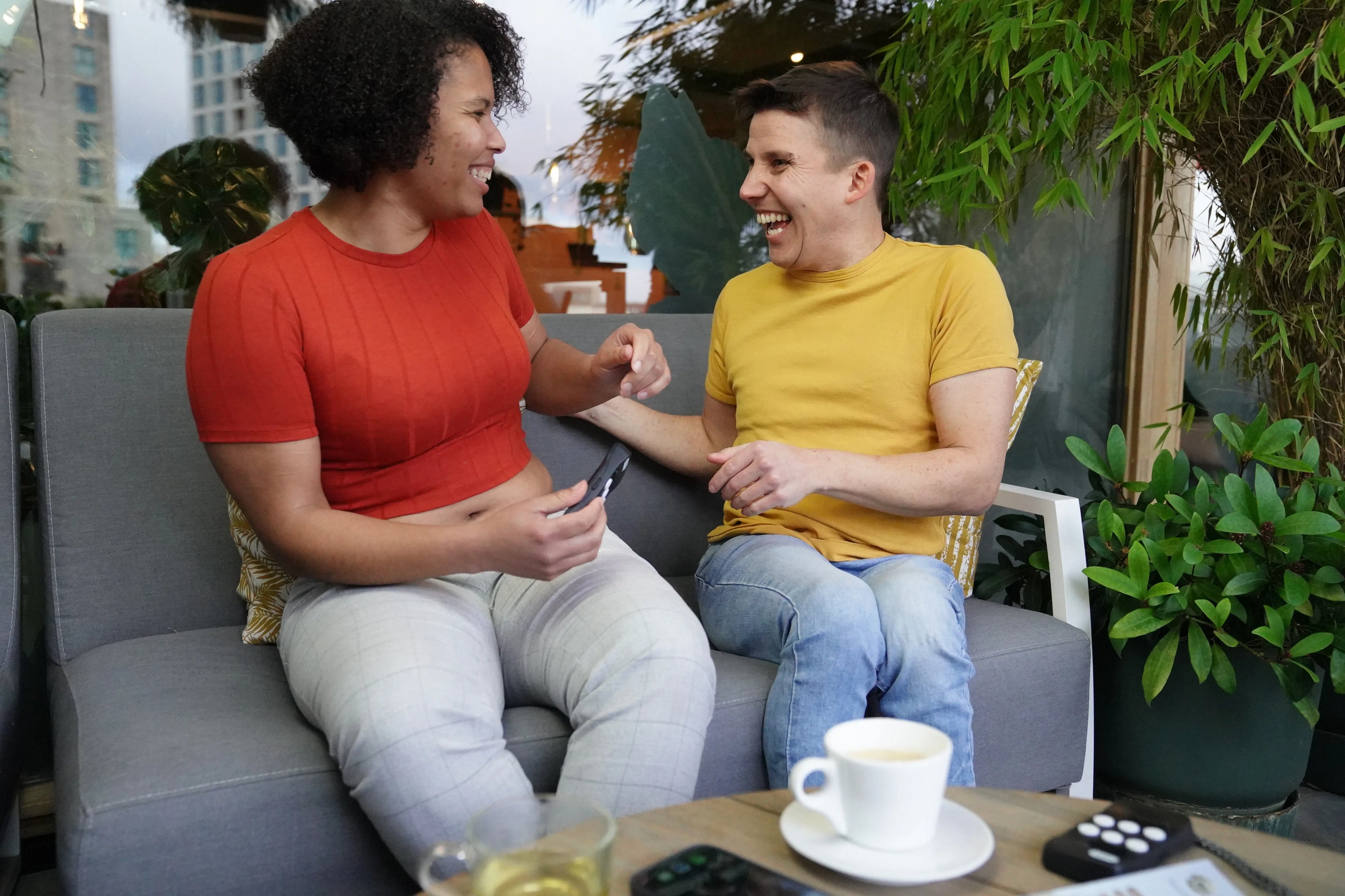 Two people sit together on a grey outdoor sofa, laughing and enjoying each other’s company. The person on the left, wearing a red top and light grey pants, holds a Hable One, a small black tactile controller with six white buttons. The person on the right, in a yellow shirt and jeans, leans in with a big smile. On the wooden table in front of them are drinks, including a cup of tea and a cup of coffee, along with another Hable One device Green plants and a softly lit background create a relax atmosphere.