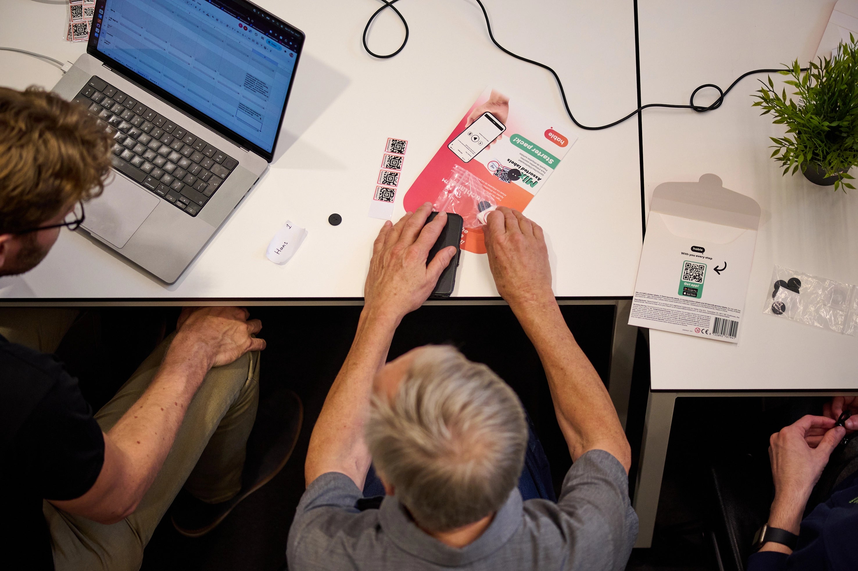A blind man uses his hands to explore a smartphone on a table during a Hable test event. In front of him are product packaging, instruction materials, and small devices. Next to him, another participant and a laptop are visible, showing a collaborative testing setup.