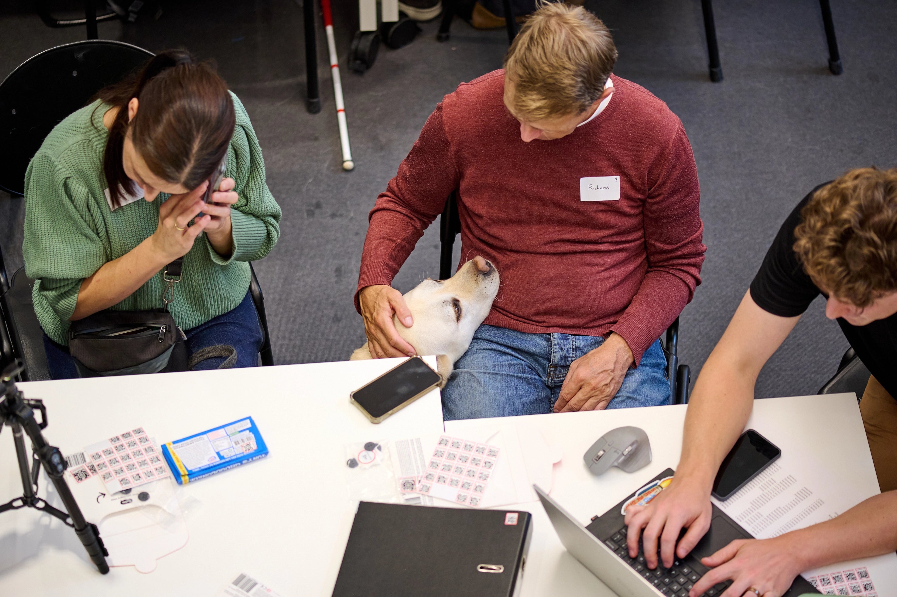 During a Hable test event, a man sits with his guide dog resting its head on his lap while he gently pets it. Next to him, a woman uses her smartphone, and across the table, another participant works on a laptop. The table is scattered with papers, stickers, and small devices.