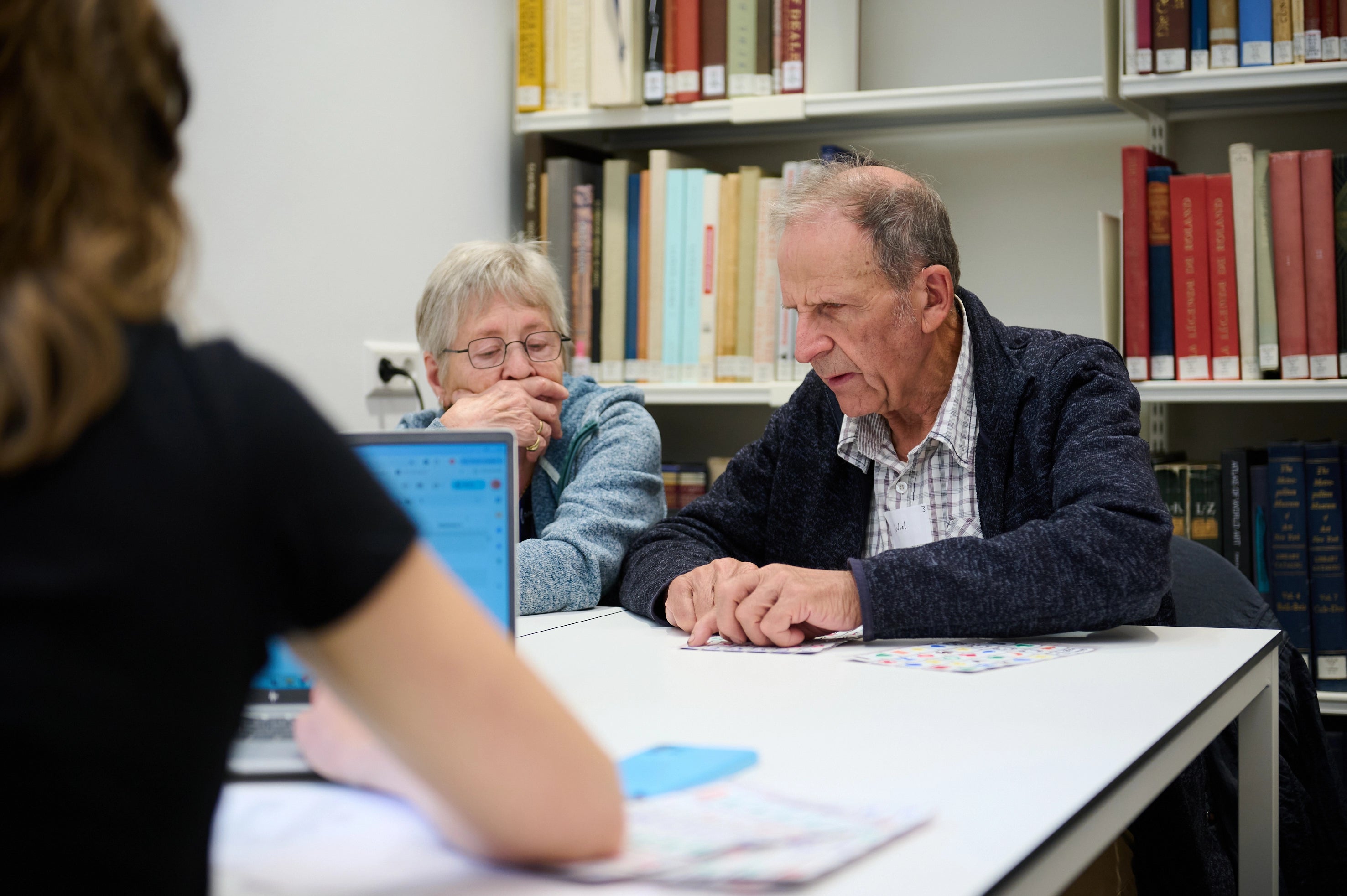 An elderly blind man sits at a table in a library, using his fingers to read a tactile sheet. Next to him, an elderly woman listens attentively with her hand resting near her face. Across the table, a facilitator with a laptop supports the session.