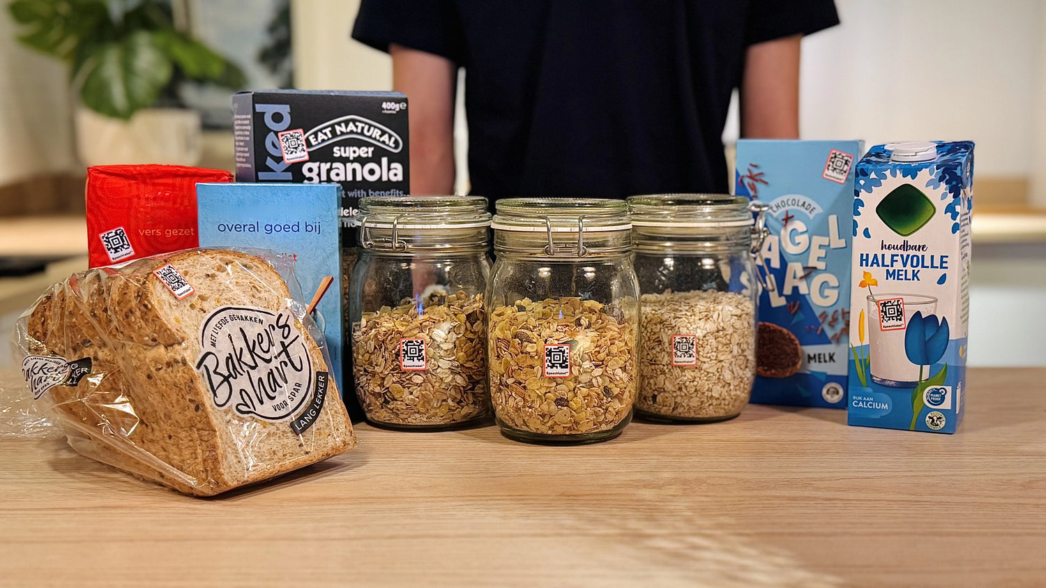 Assorted groceries displayed on a wooden table, each marked with a Speechlabel sticker. Items include a loaf of sliced bread in clear packaging, two colorful cardboard packages, three glass jars filled with different types of granola and oats, a blue carton of chocolate sprinkles, and a carton of half-fat milk. All visible stickers are Speechlabel labels designed for accessible identification with the companion app.