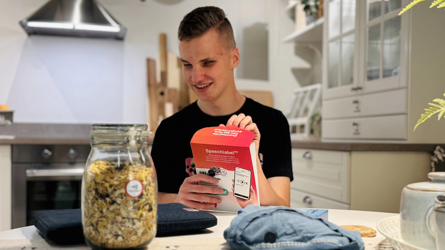 In a bright kitchen, a smiling young man opens a red-and-white Speechlabel™ starter kit box at a table. In the foreground, a glass jar of muesli has a small round Speechlabel sticker; a dark pouch, a light-blue cloth, and a teapot sit nearby. Cabinets and an oven are in the background. All labels shown are from Speechlabel, which pairs with the smartphone app pictured on the box.