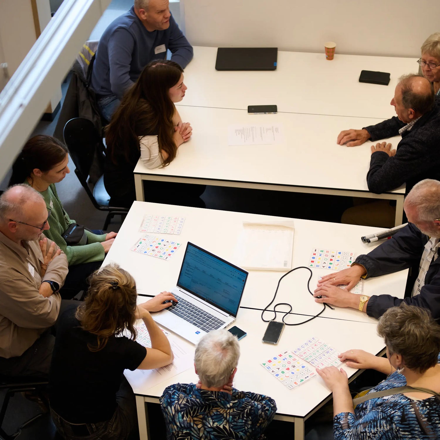 A group of people sits around tables participating in a user testing session for the Hable Easy device. Some participants use tactile button charts and a connected laptop, discussing and testing accessibility features for blind and visually impaired users.