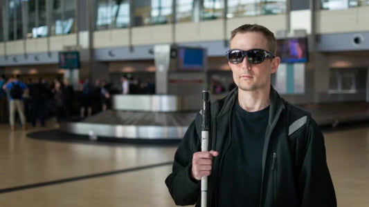 A visually impaired man standing in an airport baggage claim area holding a white cane.