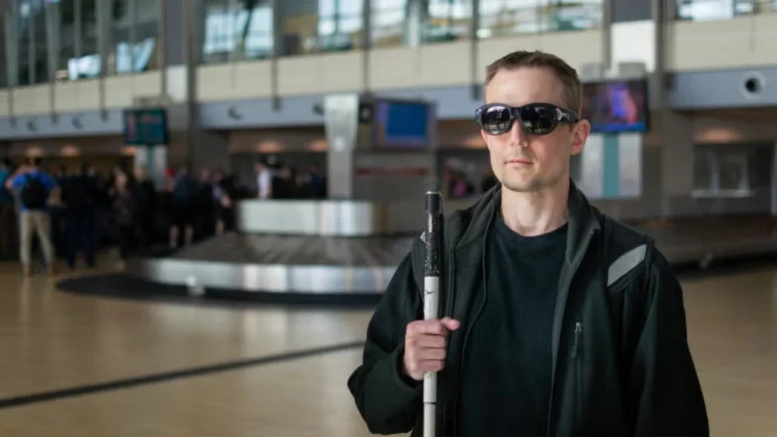 A visually impaired man standing in an airport baggage claim area holding a white cane.
