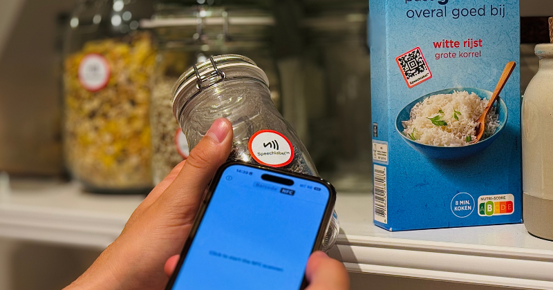 A close-up of a person holding a smartphone to scan a circular 'Speechlabel' NFC tag on a glass jar in a kitchen pantry. A box of white rice with a QR code is visible in the background.