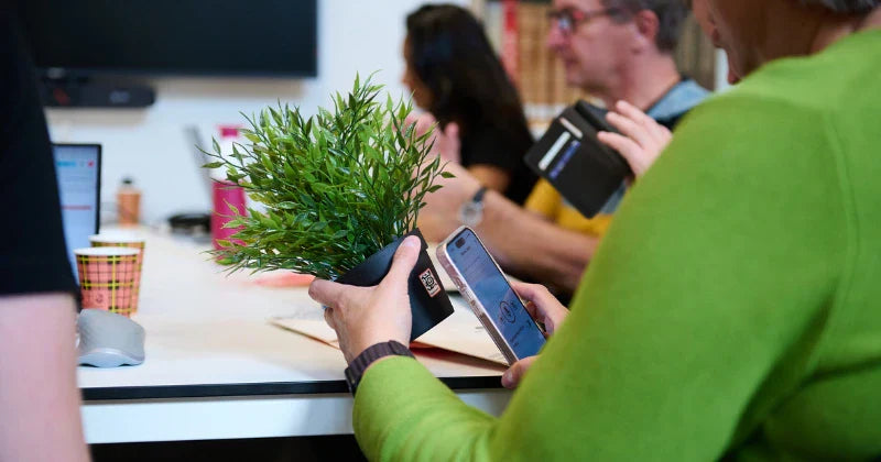 Person using the SpeechLabel app on a smartphone to scan a QR code label attached to a small potted plant during a meeting.