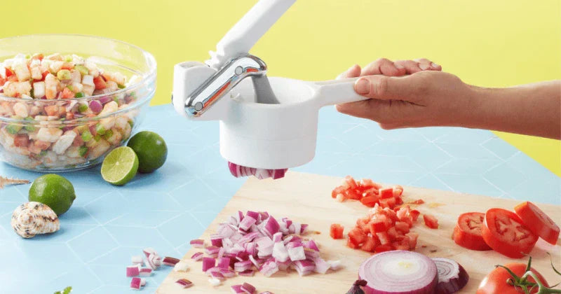 A person using a white handheld lever-style food dicer to chop a red onion over a wooden cutting board. The scene includes fresh tomatoes, limes, and bowls of shrimp ceviche on a bright blue and yellow background.