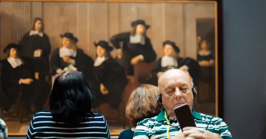 An older man with headphones sleeps while holding a smartphone in a museum gallery, positioned in front of a large, out-of-focus 17th-century style painting of several men in black robes and white collars.