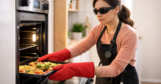 Blind woman wearing dark glasses and red oven mitts placing a tray of roasted vegetables into an open oven in a modern kitchen, with a white cane and safety device attached to her apron.