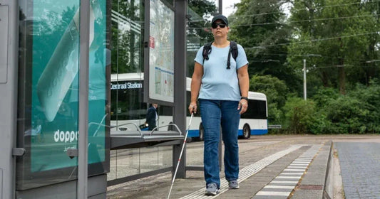 A woman with a white cane and sunglasses navigates a sidewalk using a tactile paving strip at a bus stop.
