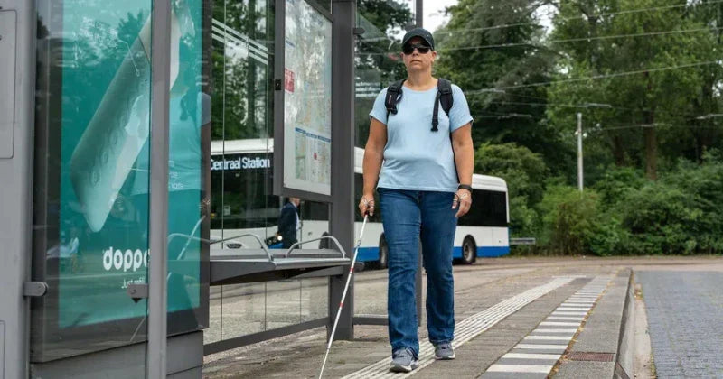A woman with a white cane and sunglasses navigates a sidewalk using a tactile paving strip at a bus stop.