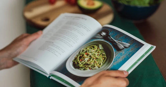 Hands holding open a cookbook to a photo of pesto pasta, with a blurred avocado and salad bowl in the background.