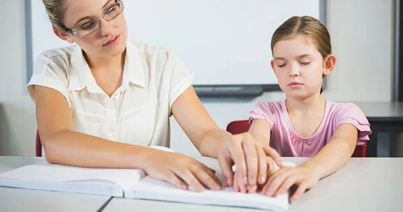  A female teacher wearing glasses helps a young girl read a Braille book by guiding her hands.