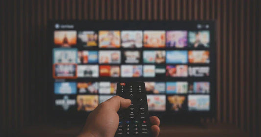 A hand holds a TV remote, pointing it towards a TV screen displaying a grid of diverse streaming content, all set against a subtly blurred wooden-paneled background.