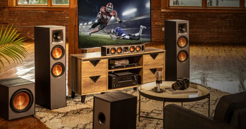A living room is set up as a home theater, featuring a large screen TV showing a football game, surrounded by a complete multi-speaker sound system on and around a wooden media console.