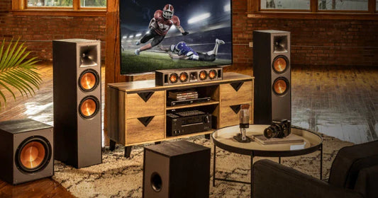 A living room is set up as a home theater, featuring a large screen TV showing a football game, surrounded by a complete multi-speaker sound system on and around a wooden media console.