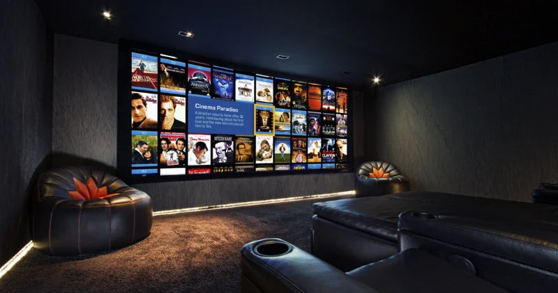 A dark, cozy home cinema features a large screen displaying movie posters and plush, comfortable seating.