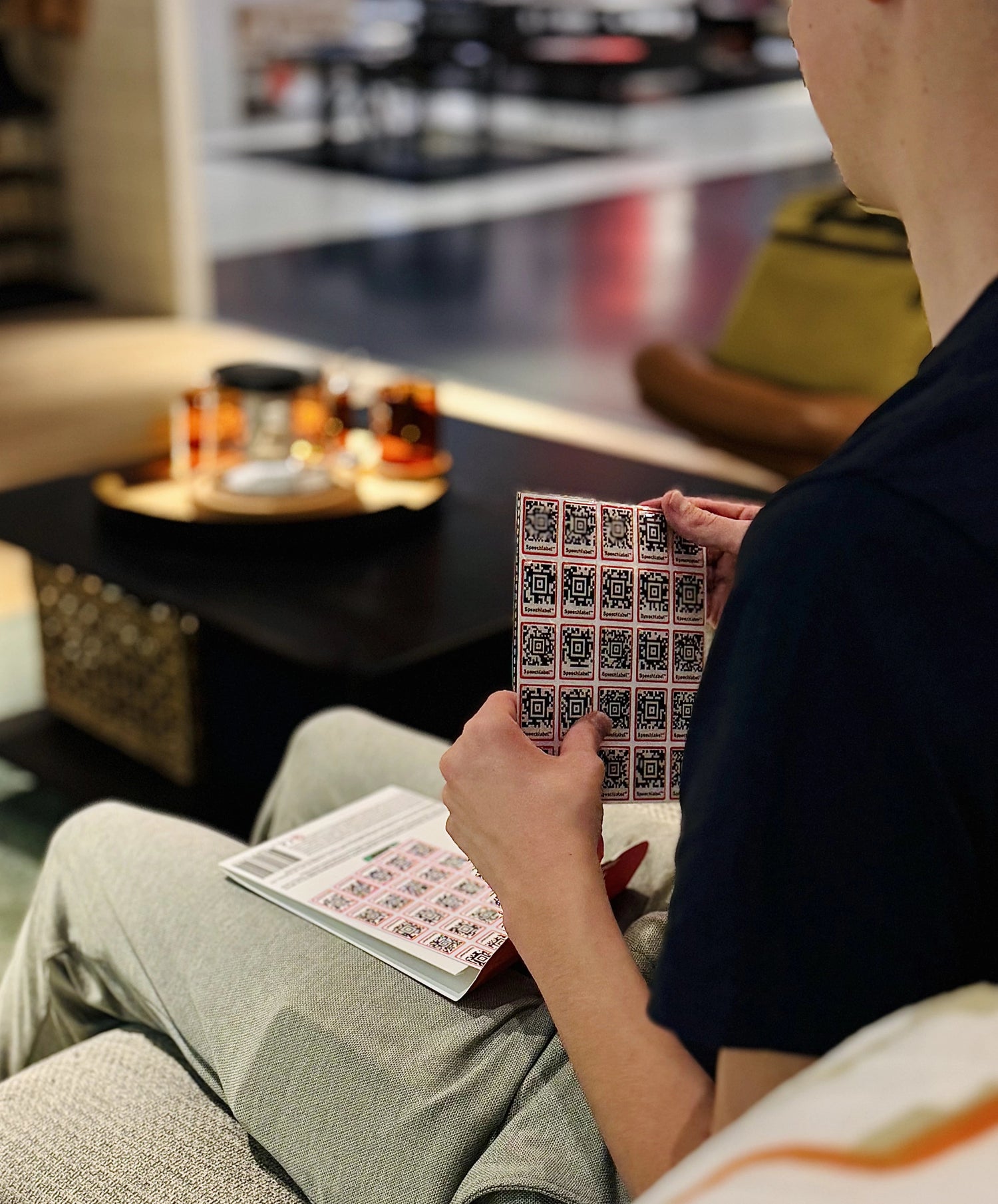 A person sitting on a sofa holds a sheet of multiple Speechlabel stickers, with another sheet resting on their lap. The stickers feature the recognizable black-and-red design used for accessible labeling. All labels shown are from Speechlabel, designed for accessible identification with the companion app.