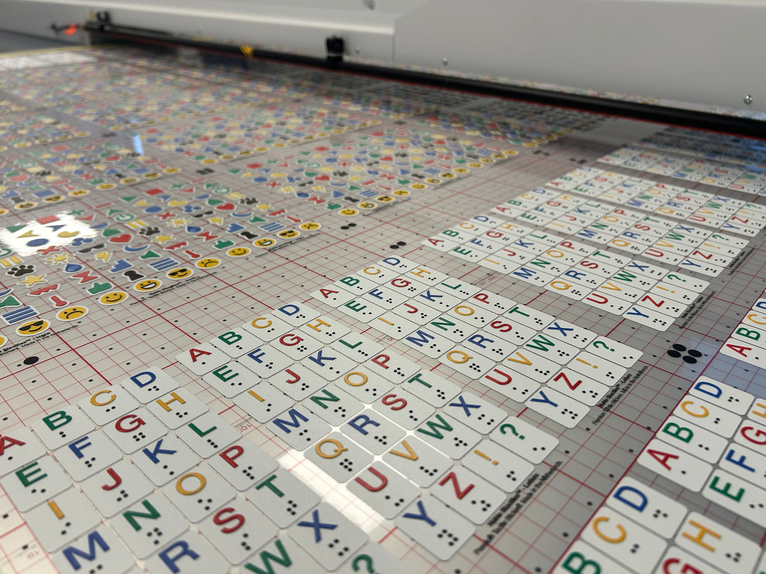 A large sheet of colorful Stactiles tactile stickers lies on a production bed, covered with rows of alphabet letters and small icons, each featuring raised Braille dots. The sheet is positioned under a printing and cutting machine, showing the manufacturing process of the tactile stickers before they are separated, coated and packaged.
