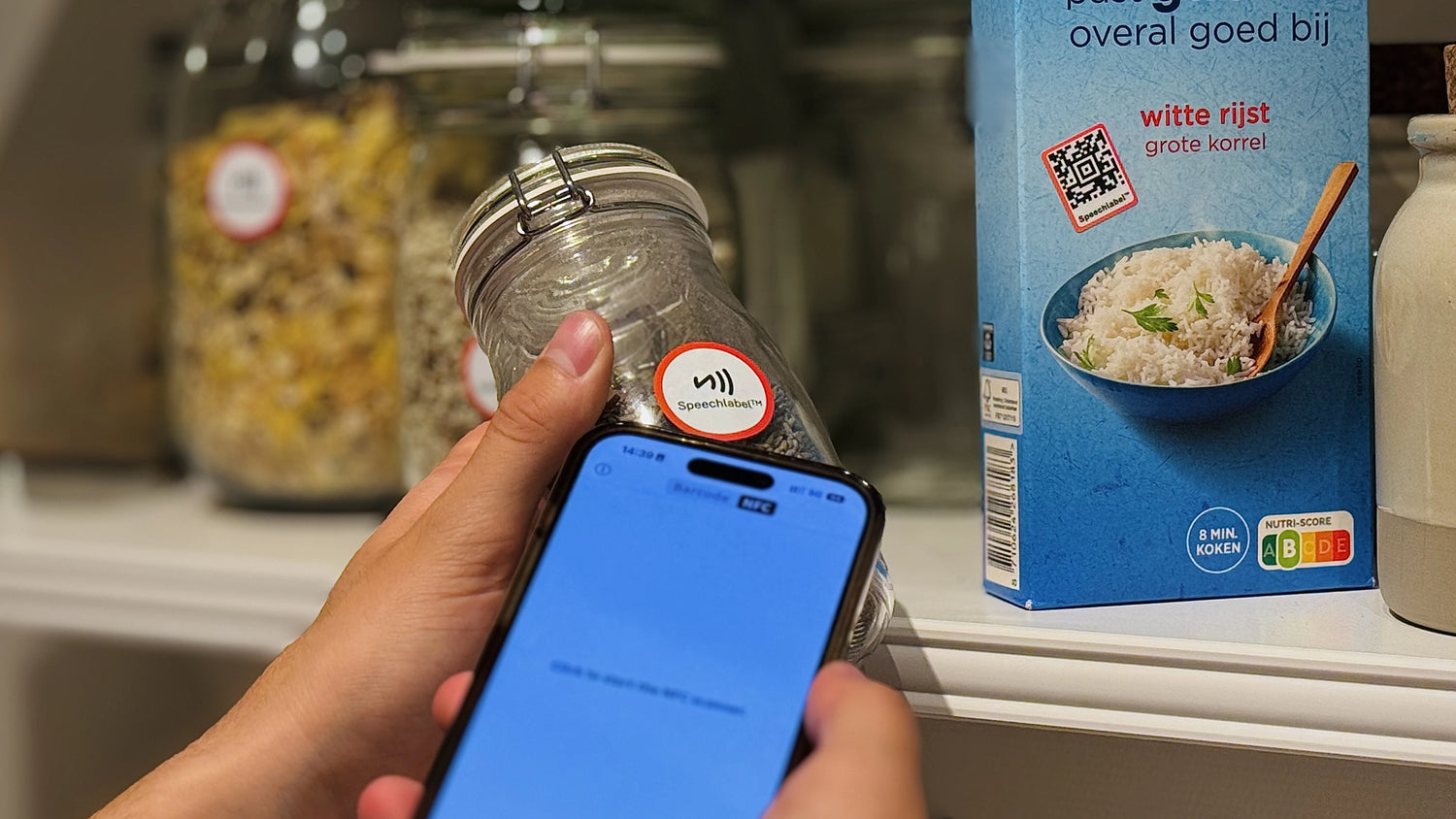 Close-up of a person holding a glass jar with a round red-and-white Speechlabel™ sticker, while scanning it with a smartphone. In the background, other labeled jars are visible along with a blue rice package also marked with a Speechlabel sticker. All labels shown are from Speechlabel, designed for accessible identification with the companion app.