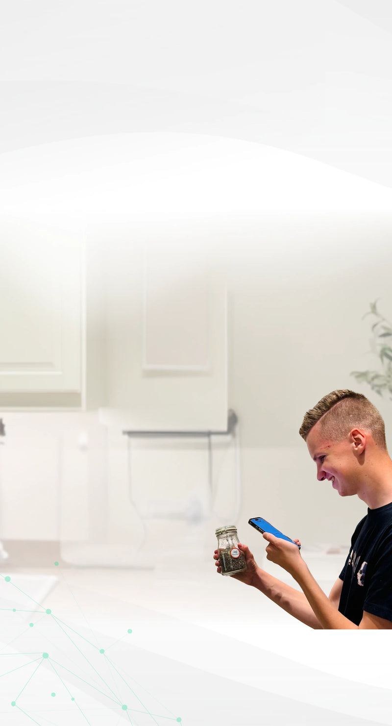 A young man smiles in a kitchen while scanning a glass jar with a round red-and-white Speechlabel™ sticker using his smartphone. A speech bubble shows the audio output: ‘Black Tea, best before: 31/12/1999.’ The scene illustrates how Speechlabel stickers make everyday items easy to identify.