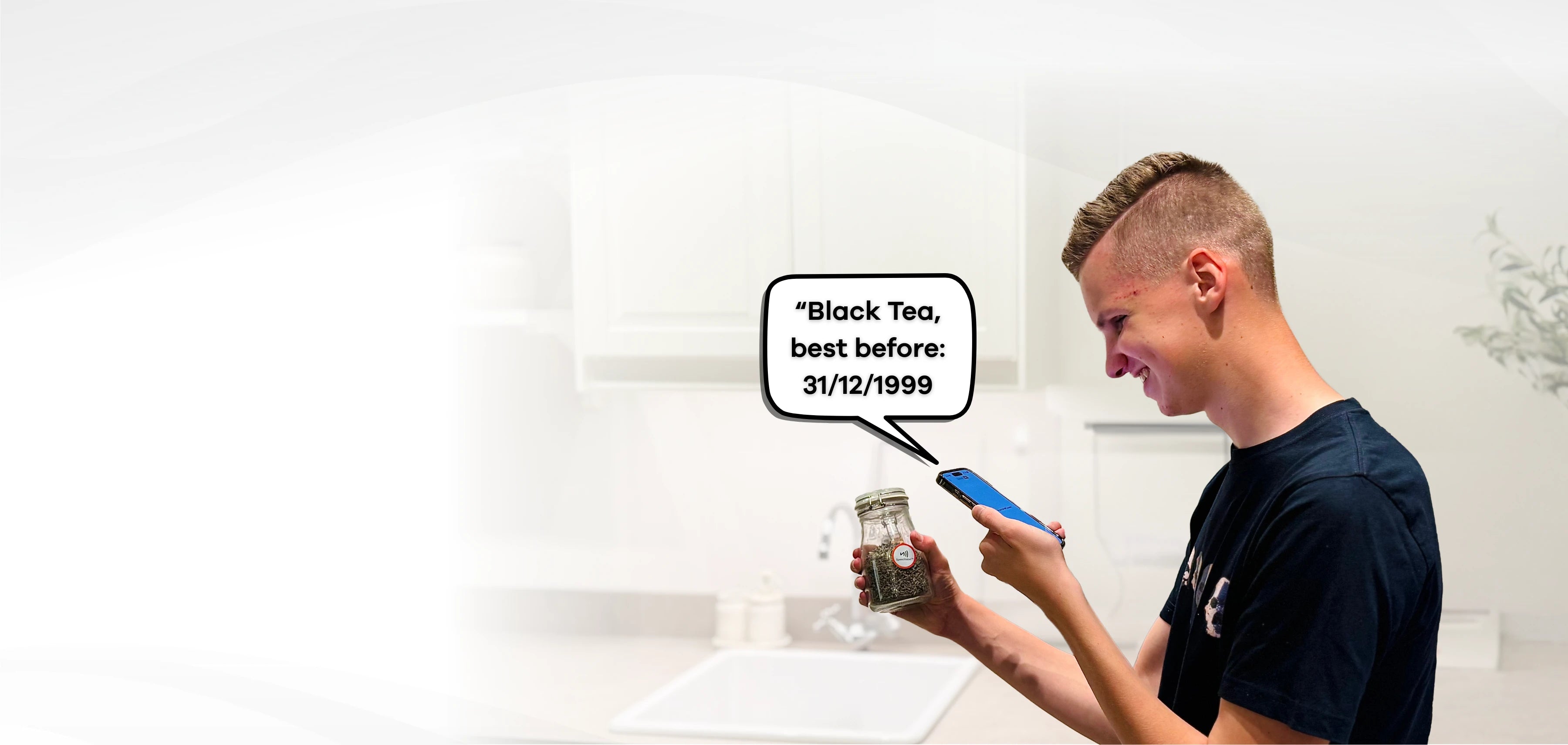 A young man smiles in a kitchen while scanning a glass jar with a round red-and-white Speechlabel™ sticker using his smartphone. A speech bubble shows the audio output: ‘Black Tea, best before: 31/12/1999.’ The scene illustrates how Speechlabel stickers make everyday items easy to identify