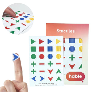 A composite image showcasing the Stactiles Basic Shapes set. A circular inset in the top-left shows fingers feeling different textured stickers. In the bottom-left, a blue textured chevron sticker is shown on a fingertip for scale. The center features a full sheet of 24 stickers in green, red, blue, and yellow. Shapes include triangles, squares, circles, pluses, minuses, and chevrons, many of which feature unique tactile patterns like vertical ridges, cross-hatching, and wavy lines.