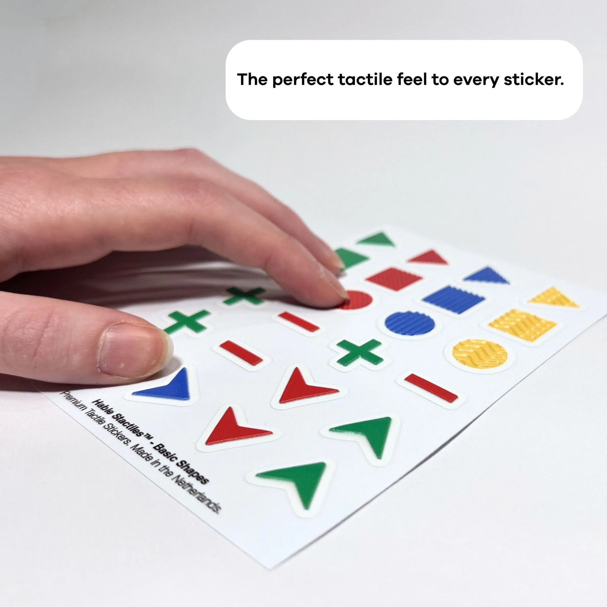 A close-up, angled photograph of a hand scanning a sheet of basic shape stickers. The focus is on the diverse surface textures: a red square with a grid pattern, a blue circle with vertical ridges, and a yellow circle with a wavy embossed surface. These textures provide additional tactile cues beyond the outer shape. Text at the bottom reads: "The perfect tactile feel to every sticker."