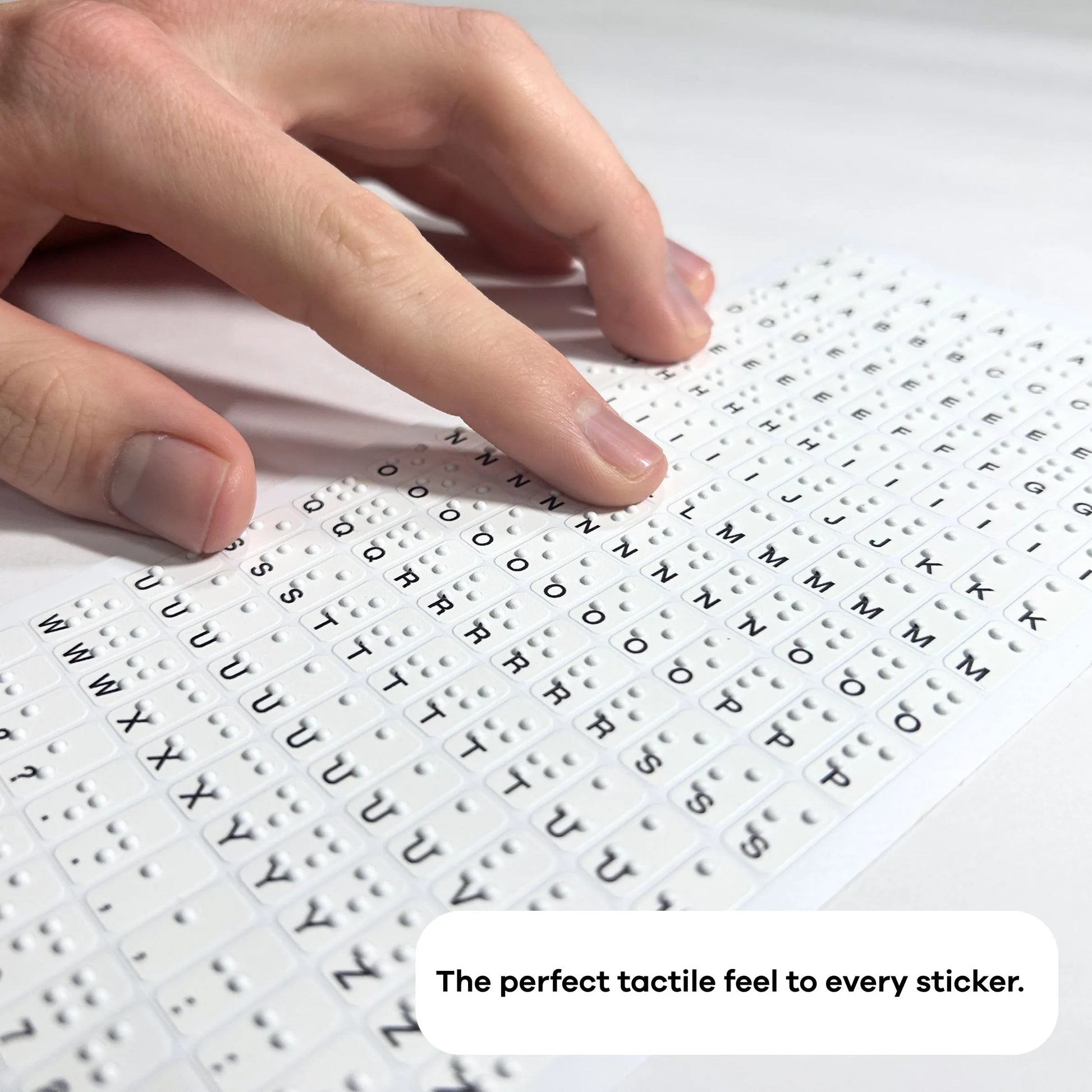 A close-up, angled photograph of a person’s hand moving across a full sheet of small Braille stickers. Each white sticker features a small black printed letter and raised tactile dots. The focus emphasizes the high-quality 3D texture, with a text overlay at the bottom: "The perfect tactile feel to every sticker."