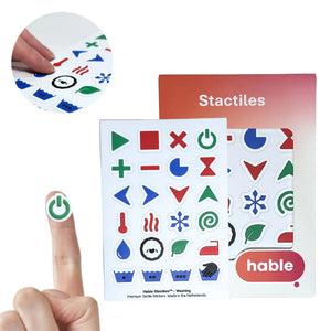 A composite image of the Hable Stactiles Washing set. A circular inset shows fingers touching laundry-specific symbols. In the bottom-left, a green power symbol sticker is shown on a fingertip for scale. The full sheet of 24 stickers includes specialized icons such as an iron, a sheep (wool cycle), a snowflake (cold wash), heat waves, a thermometer, and water tubs with embossed dots representing different temperatures. The Hable packaging is visible on the right.