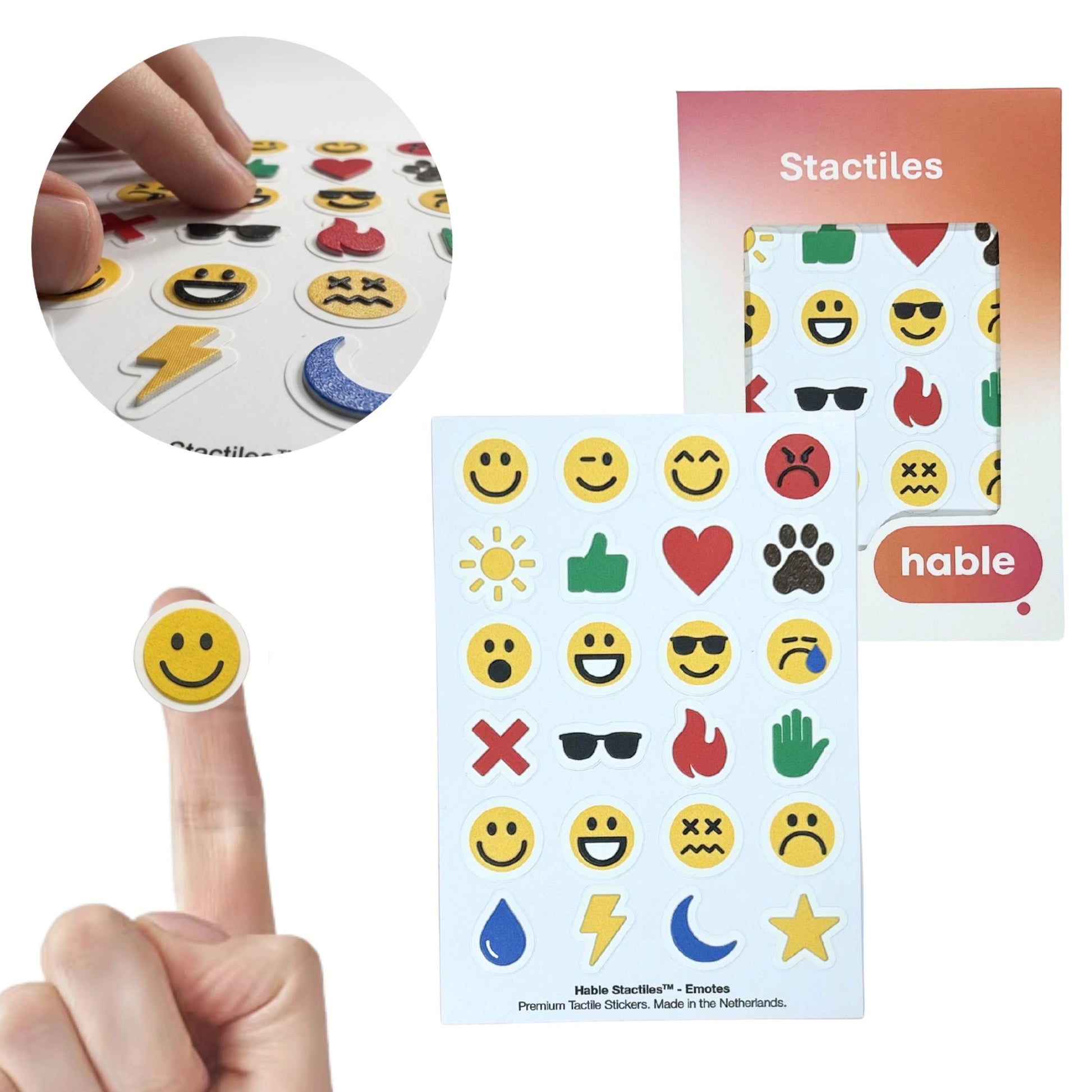 A composite image showcasing the Stactiles Emoji set. A circular inset in the top-left shows fingers exploring a sheet of colorful, raised emoji stickers. In the bottom-left, a classic yellow smiley face sticker is shown on a fingertip for scale. The main image features a sheet of 24 unique haptic stickers—including various facial expressions, a heart, a fire symbol, and a thumbs up—displayed next to the Hable retail packaging.