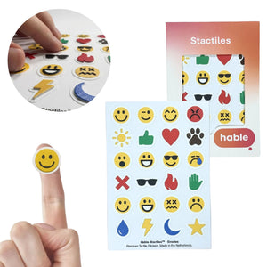 A composite image showcasing the Stactiles Emoji set. A circular inset in the top-left shows fingers exploring a sheet of colorful, raised emoji stickers. In the bottom-left, a classic yellow smiley face sticker is shown on a fingertip for scale. The main image features a sheet of 24 unique haptic stickers—including various facial expressions, a heart, a fire symbol, and a thumbs up—displayed next to the Hable retail packaging.