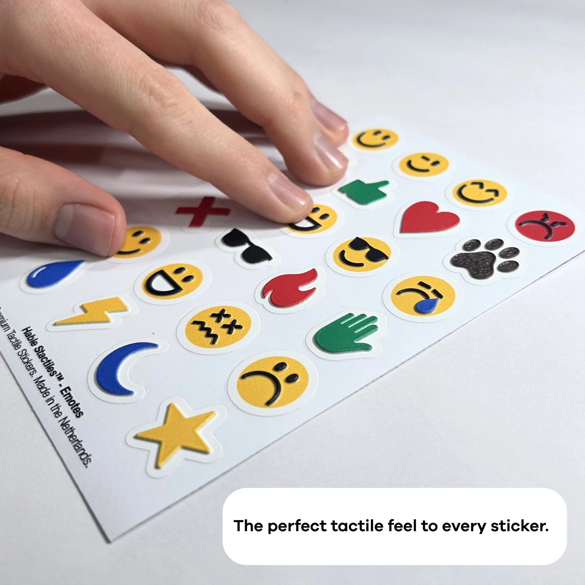 A close-up, angled photograph of a person’s hand scanning a sheet of Stactiles Emojis. The focus is on the significant 3D height and detailed texture of the icons, such as the ridges on the lightning bolt and the embossed facial features of the smiley faces. A text overlay at the bottom reads: "The perfect tactile feel to every sticker."