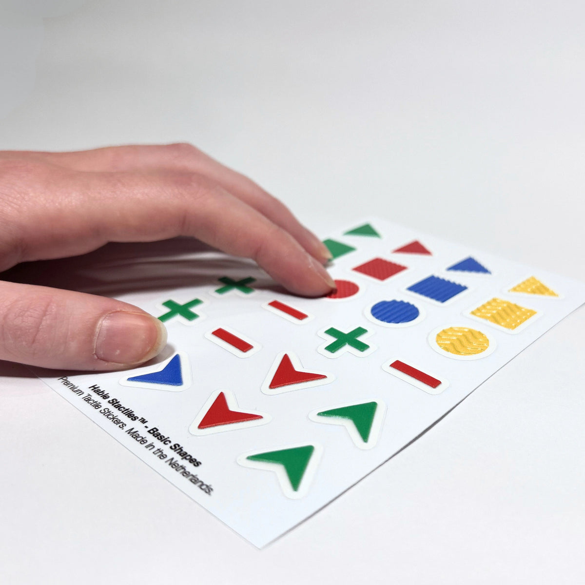 Perspective view of a hand touching the raised textured surface of the geometric shapes on the "Hable Stactiles - Basic Shapes" sticker sheet.