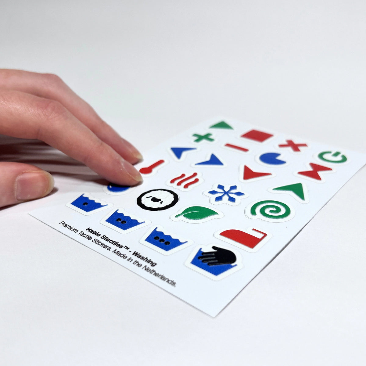 Angled perspective photo of a hand feeling the raised laundry cycle icons and water level symbols on the "Hable Stactiles - Washing" sticker sheet.