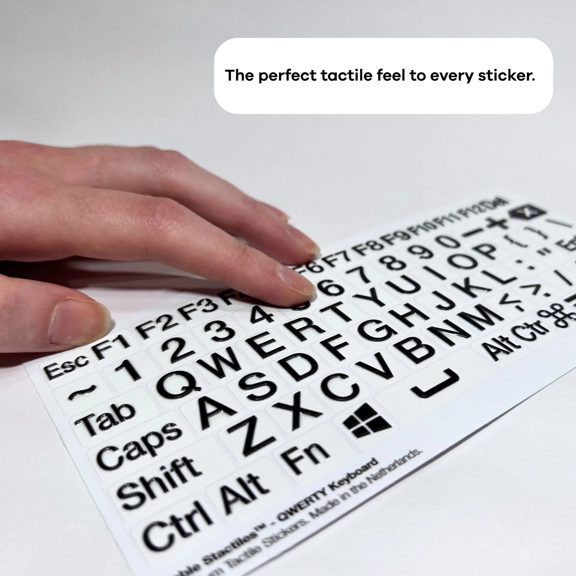 A close-up, angled photograph of a person's hand moving across a sheet of QWERTY keyboard stickers. The shot highlights the significant 3D height of the black letters and symbols, which provides clear tactile feedback for touch typing. A text overlay at the top reads: "The perfect tactile feel to every sticker."