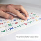 A close-up, angled photograph focused on the texture of the alphabet stickers. A person's fingers are shown touching the raised, colorful letters and the corresponding black Braille dots underneath them on a white sheet. A text overlay at the bottom reads, "The perfect tactile feel to every sticker."