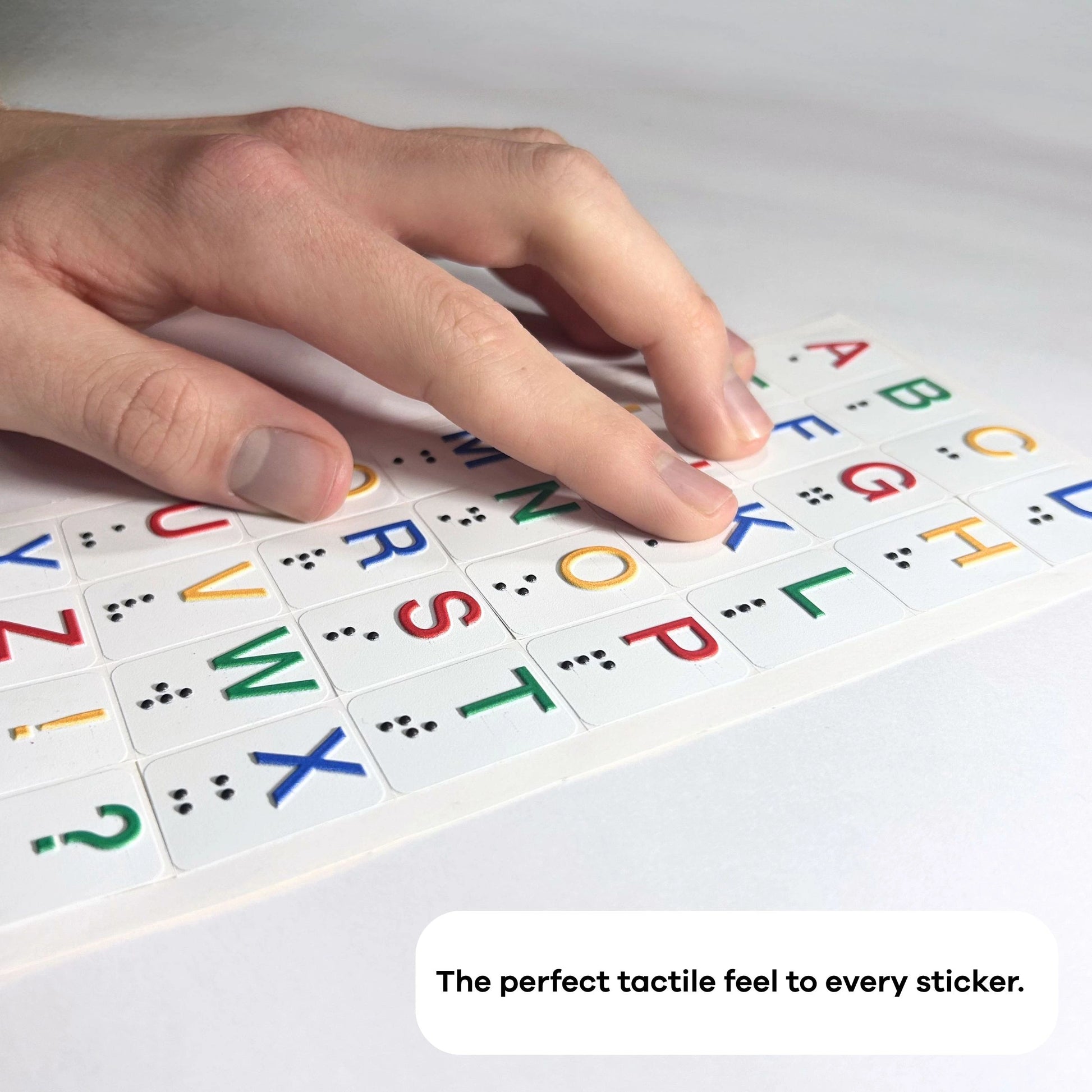 A close-up, angled photograph focused on the texture of the alphabet stickers. A person's fingers are shown touching the raised, colorful letters and the corresponding black Braille dots underneath them on a white sheet. A text overlay at the bottom reads, "The perfect tactile feel to every sticker."
