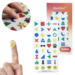 A composite image featuring fingers touching various colorful 3D tactile stickers on a white sheet. The icons include a red heart, a yellow star, and a blue crescent moon. In the bottom-left corner, a yellow lightning bolt sticker is shown placed on a person’s fingertip to demonstrate its size. The Hable Stactiles product packaging and a full sheet of icons are displayed on the right.
