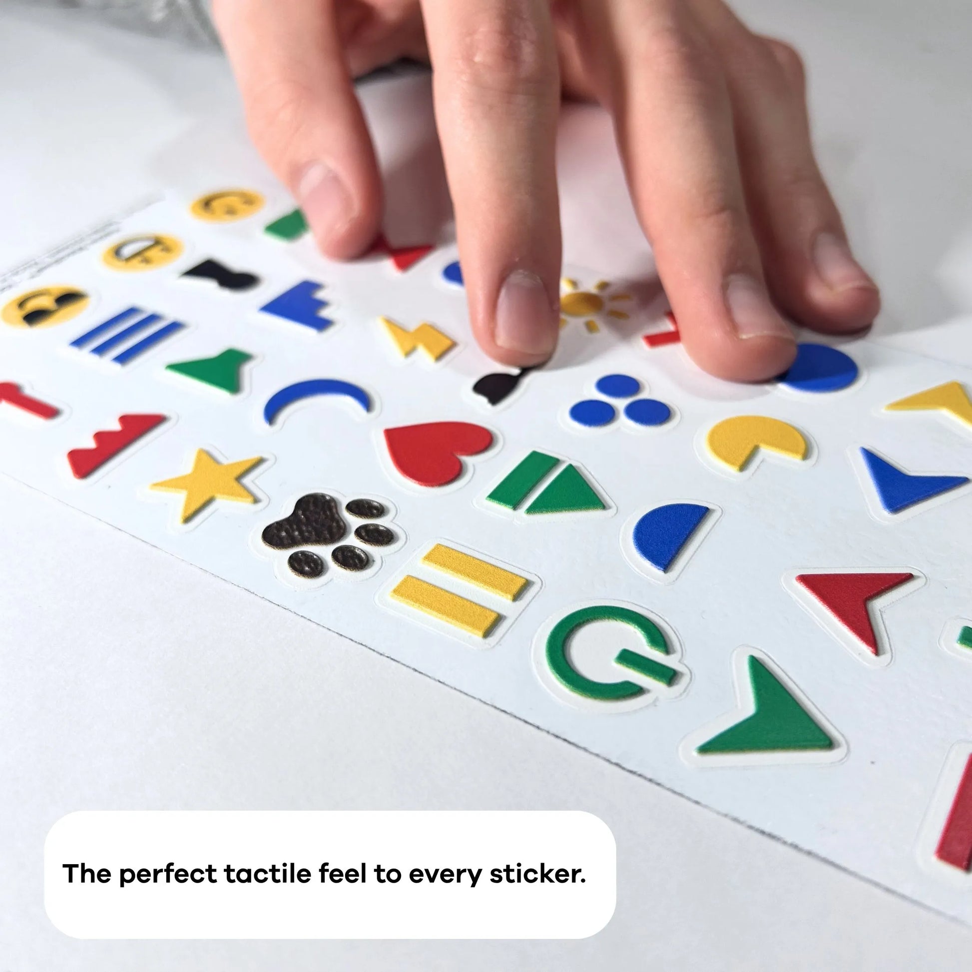 A close-up, angled photograph of a hand moving across a sheet of raised, colorful tactile stickers. The focus is on the 3D texture of the icons, such as a green power symbol and a black paw print. A text overlay at the bottom reads, "The perfect tactile feel to every sticker."