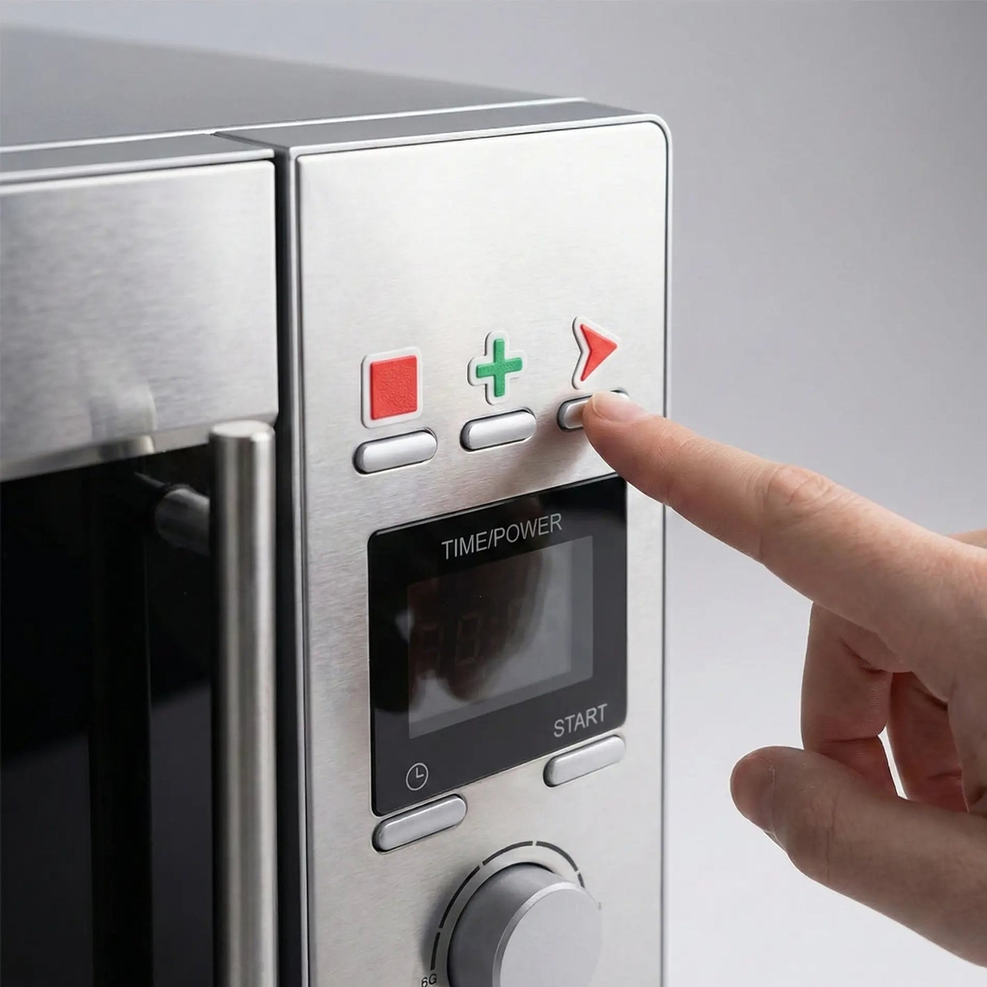 A close-up photograph of a stainless steel microwave control panel, modified with colorful tactile stickers for accessibility. A red square sticker, a green plus sign sticker, and a red triangle arrow sticker are placed above three distinct buttons. A person's index finger is pressing the button directly beneath the raised red arrow sticker, demonstrating how the tactile markers help locate specific functions by touch.