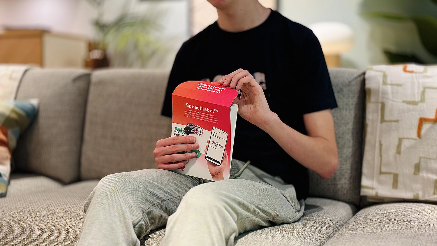 A person sitting on a couch opens a red-and-white Speechlabel™ starter kit box, which features an image of a smartphone with the Speechlabel app displayed. Cushions and a patterned throw are visible on the sofa. All labels and packaging shown are from Speechlabel, designed for accessible identification with the companion app.