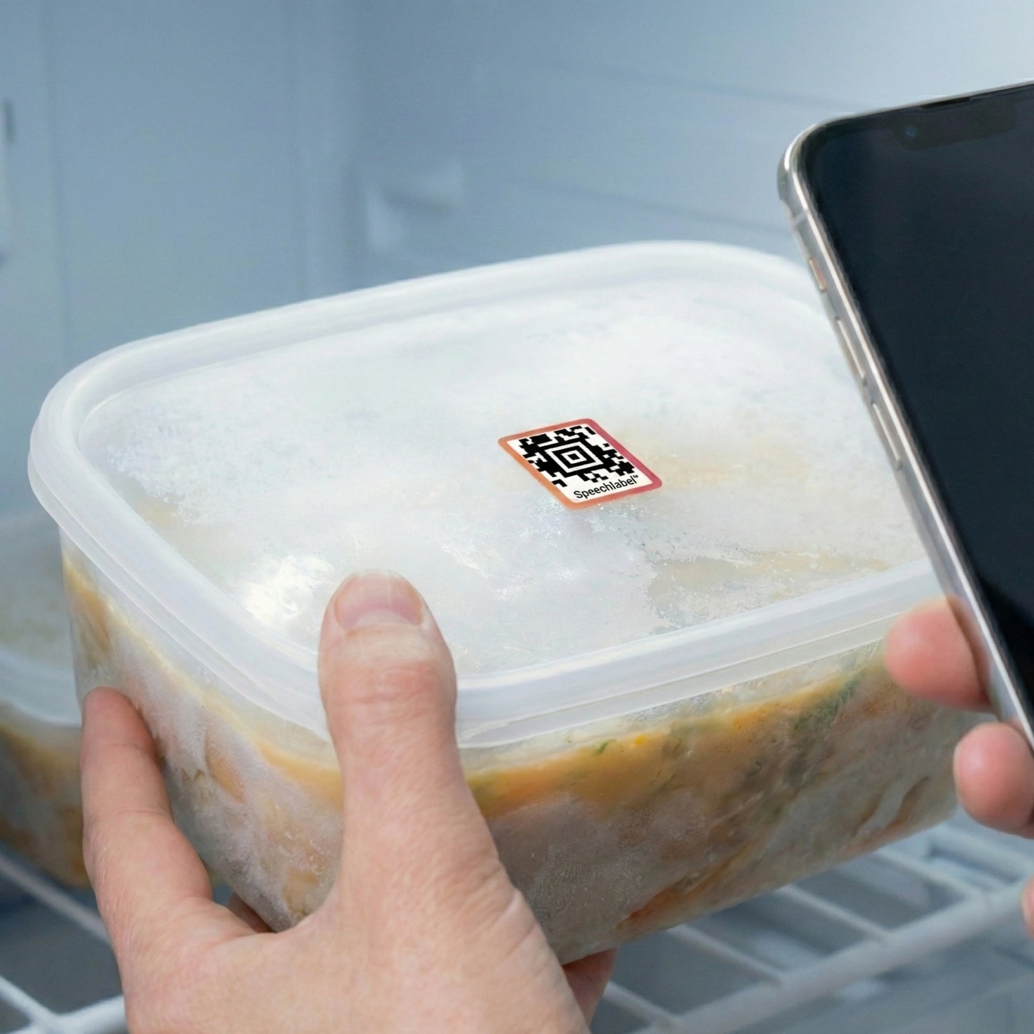 A hand holding a frosted food container in a freezer with a thin SpeechLabel QR code sticker on the lid, while a smartphone is positioned to scan it.