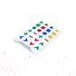 An angled perspective view of a white sheet of "Hable Stactiles - Basic Shapes" tactile stickers. This viewpoint emphasizes the raised, three-dimensional profile of the various colorful shapes, including triangles, squares, circles, plus signs, minus signs, and chevrons. The stickers are displayed in green, red, blue, and yellow, each featuring distinct tactile patterns such as smooth surfaces, dots, vertical stripes, and wavy lines. Product text is visible at the bottom left corner of the sheet.