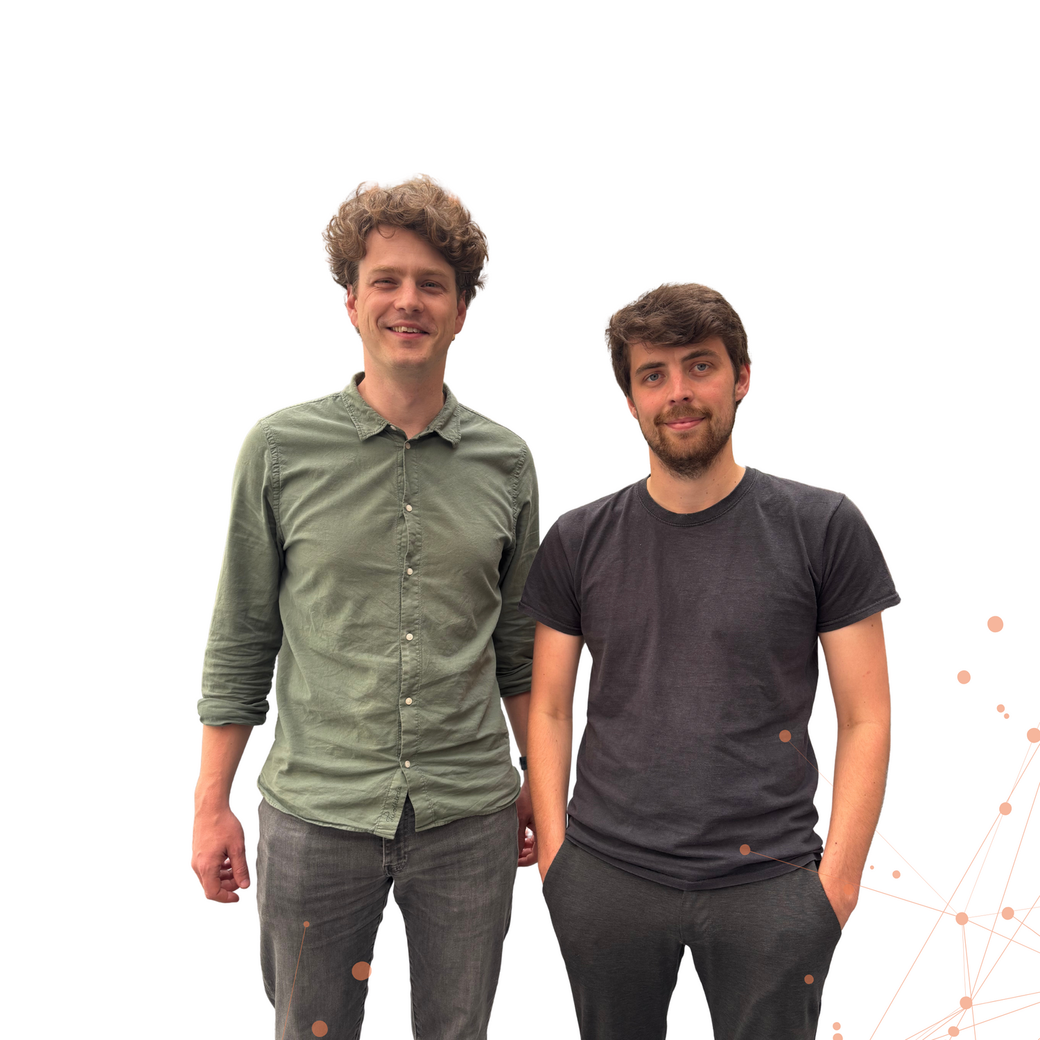 Two men standing side by side and smiling in front of a white background with a faint network graphic in the bottom right corner. The man on the left has curly hair and is wearing a green button-up shirt with grey jeans, while the man on the right has dark hair and a beard, wearing a black t-shirt and dark pants.