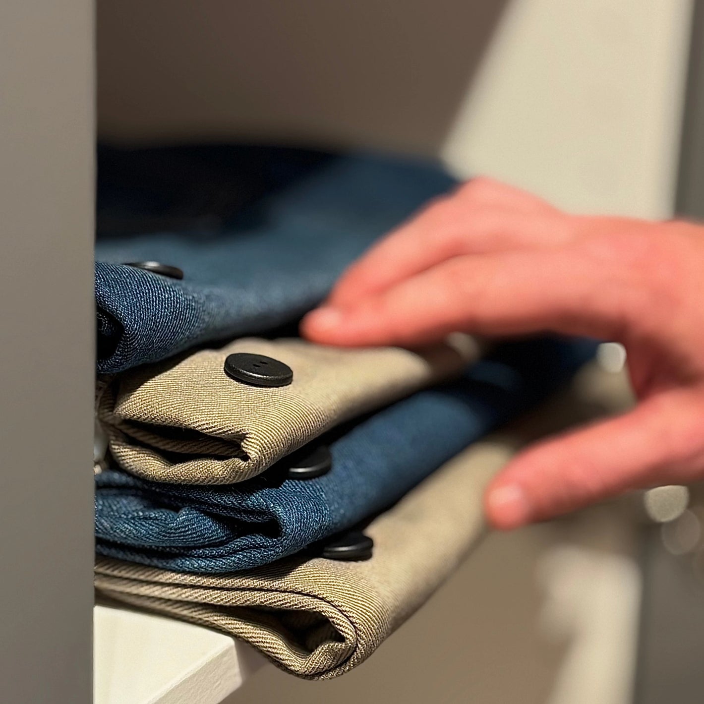 Close-up of a person’s hand touching neatly folded clothing on a shelf. The garments, in shades of blue and beige, are fastened with large black buttons. All items are prepared for use with Speechlabel, designed for accessible identification with the companion app.